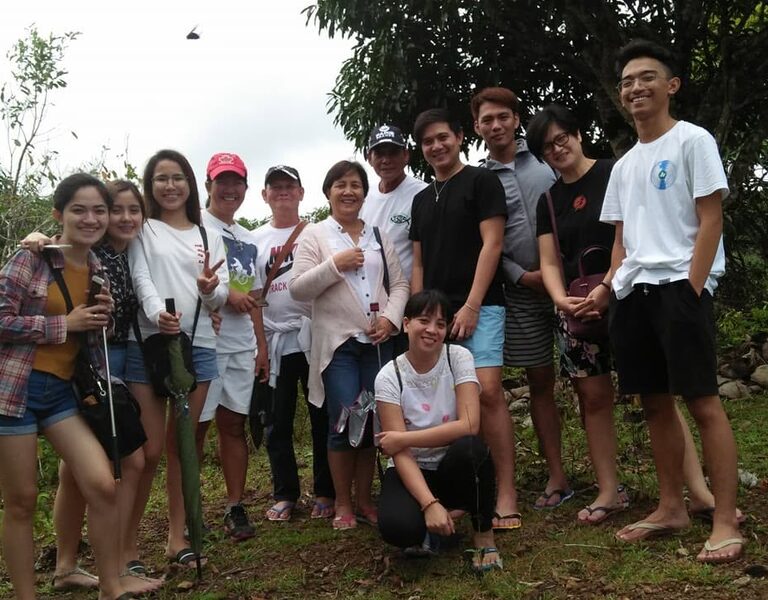 About a dozen people of all ages posed together on cleared ground, a young tree in its planting bag staked in front of them